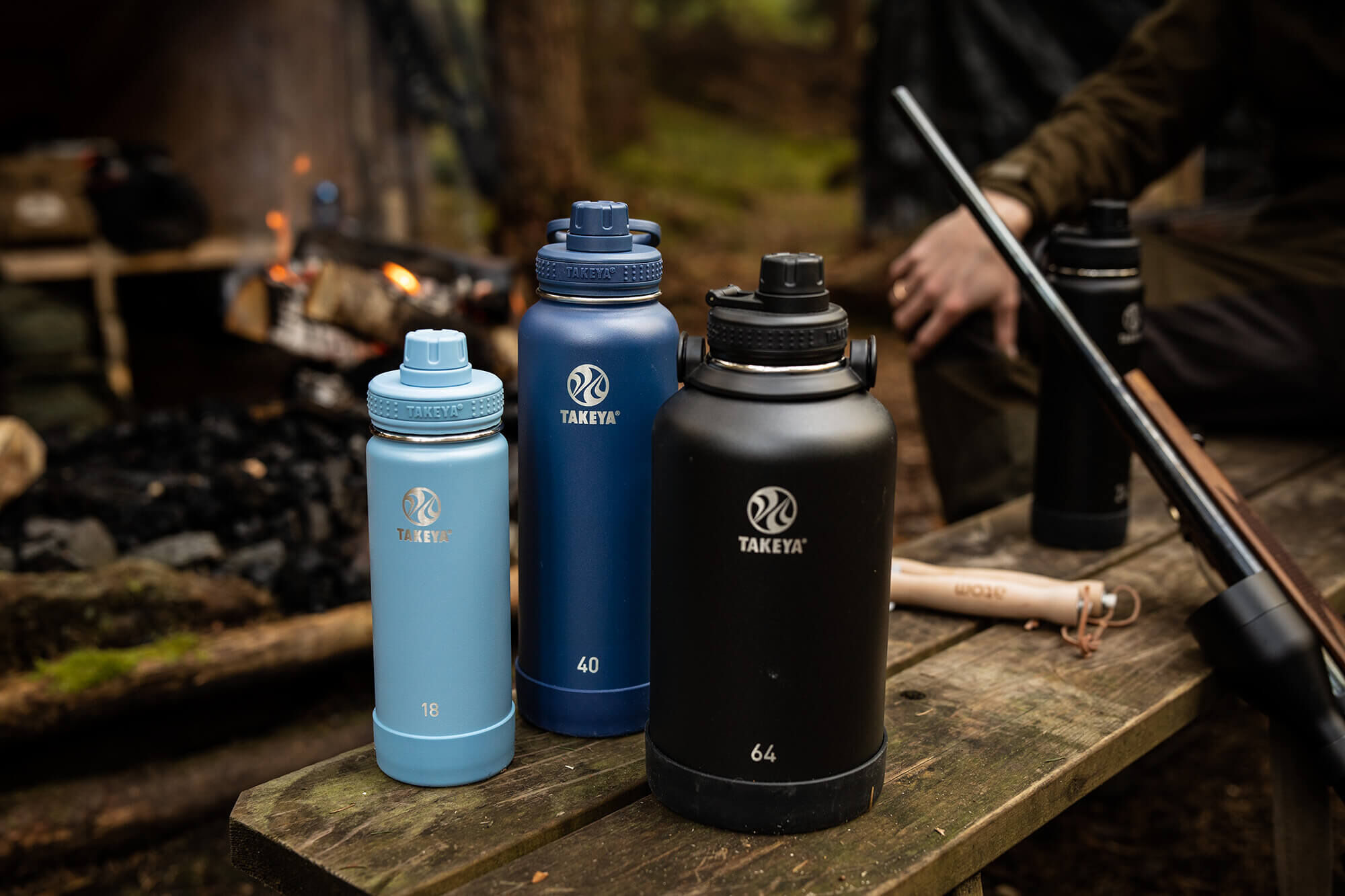 Drei Takeya Isolierflaschen in Blau und Schwarz auf einem Holztisch beim Camping, mit Lagerfeuer im Hintergrund.