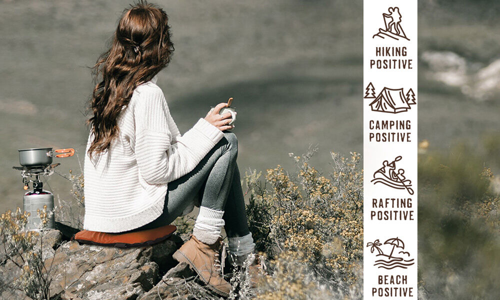 Frau genießt Outdoor-Aktivitäten in der Natur Junge Frau sitzt auf einem Stein in der Natur, hält eine Tasse, neben ihr ein Campingkocher. Symbole für Hiking, Camping, Rafting und Beach-Aktivitäten.