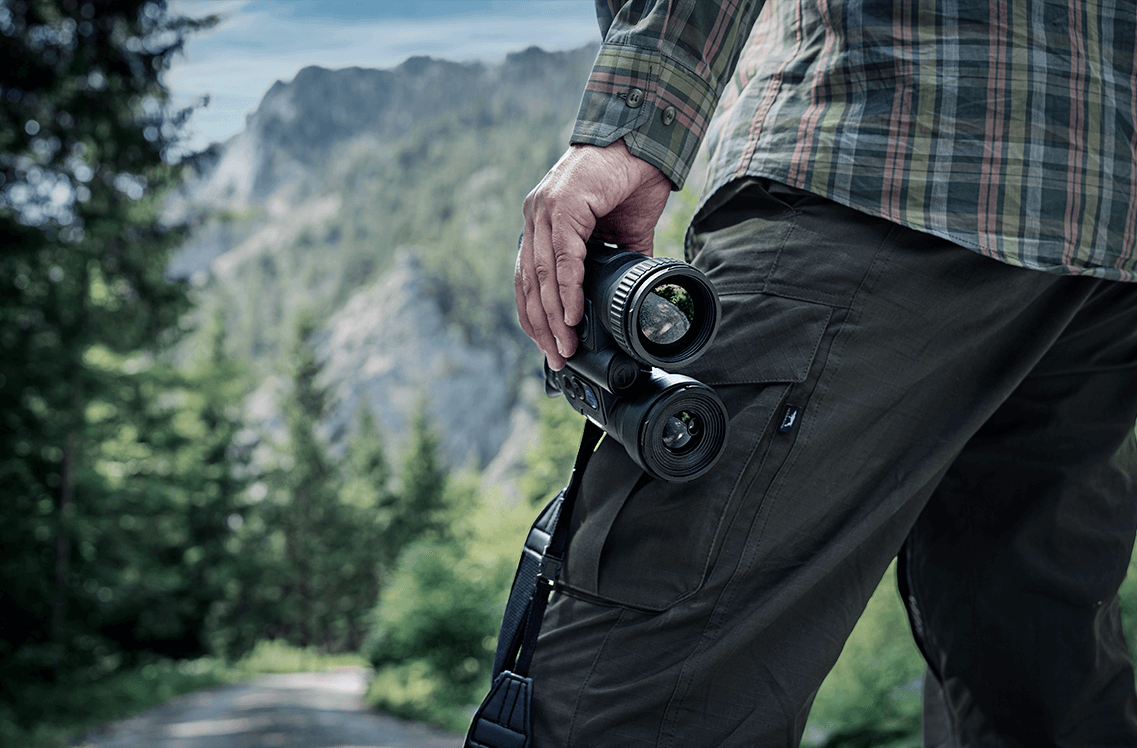 Ein Mann in Karohemd mit Fernglas in der Hand steht in einer bergigen und bewaldeten Umgebung.