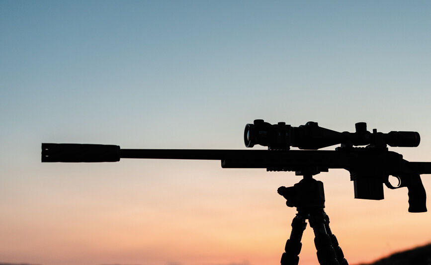 Das Bild zeigt die Silhouette eines Gewehrs, das auf einem Stativ steht, vor einem farbenfrohen Abendhimmel.