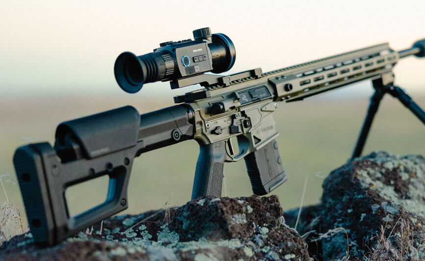 Ein taktisches Gewehr mit montiertem Zielfernrohr und Zweibein steht auf Felsen in einer ländlichen Umgebung.